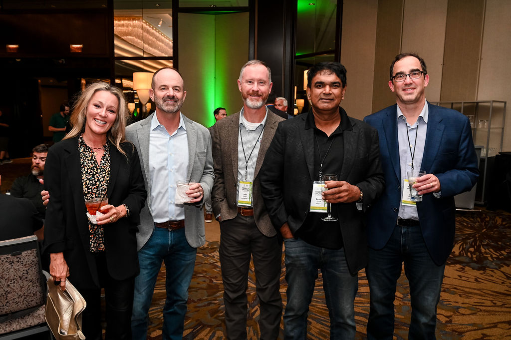 Five adults wearing conference badges stand together at an indoor networking event, smiling and holding drinks.