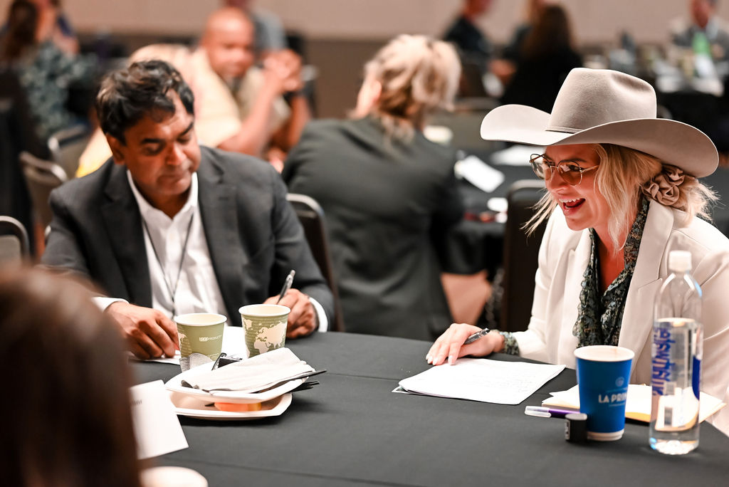 Two attendees sit at a round table during a conference session, reviewing papers and taking notes.