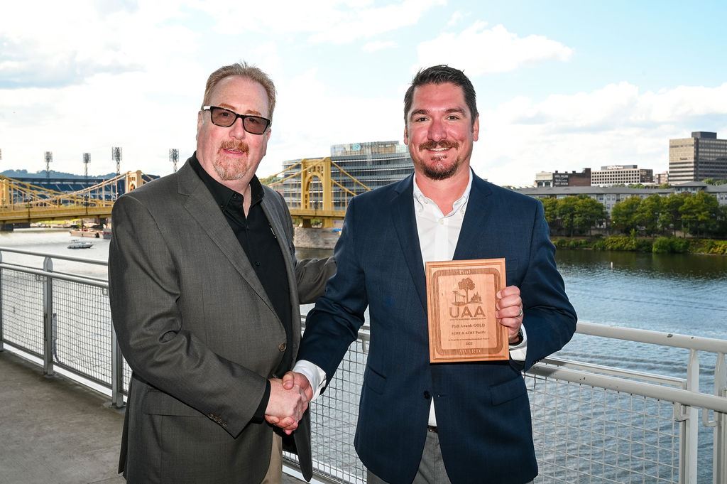 Two men in business attire shaking hands outdoors on a riverfront with bridges in the background; one is holding a wooden award plaque.
