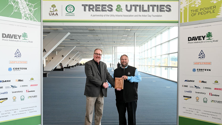 Two men shake hands and hold an award plaque beneath a “Trees & Utilities” event sign.