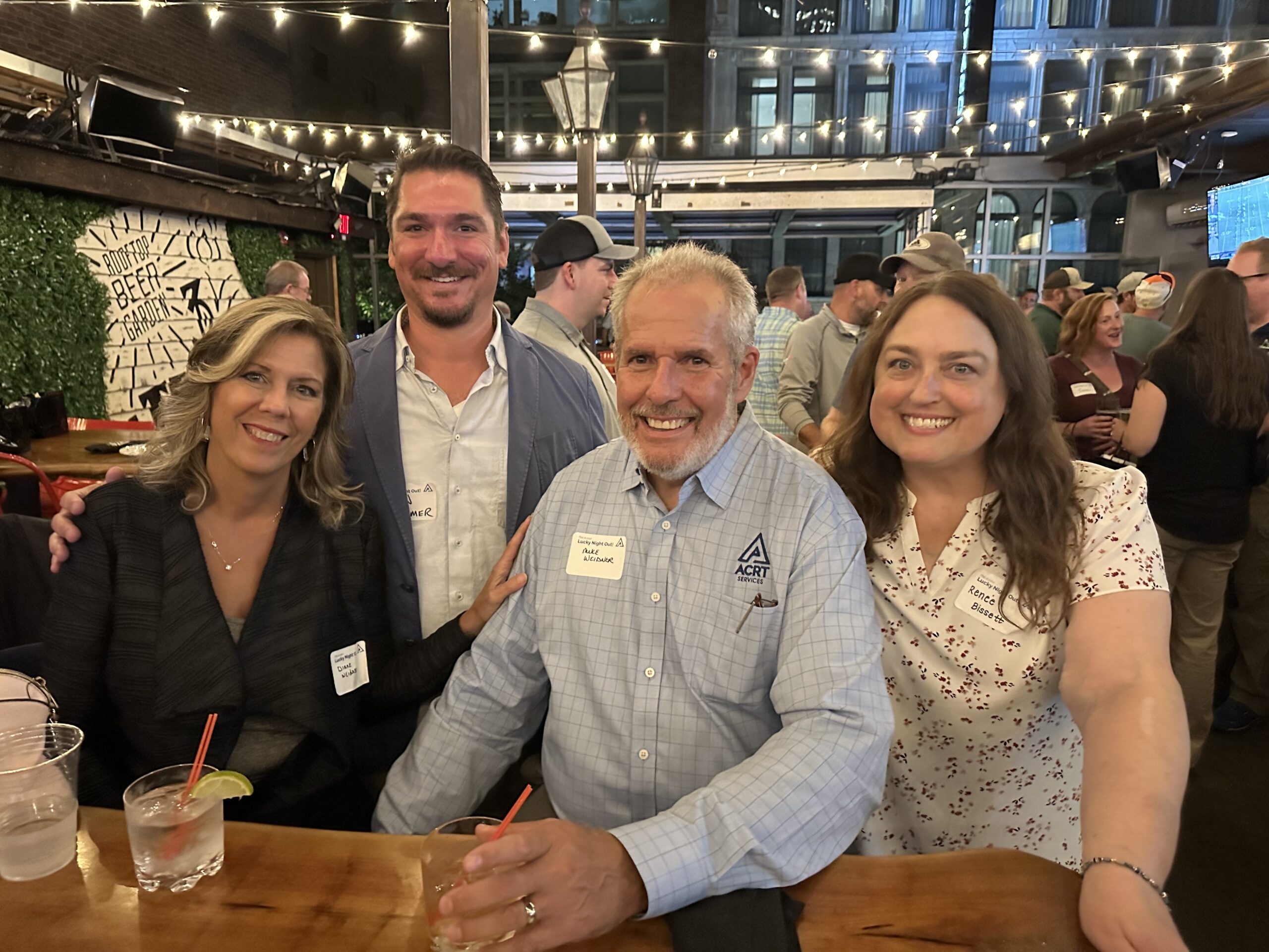 Four attendees smile together at a bar during a networking event, with drinks on the counter and string lights overhead.