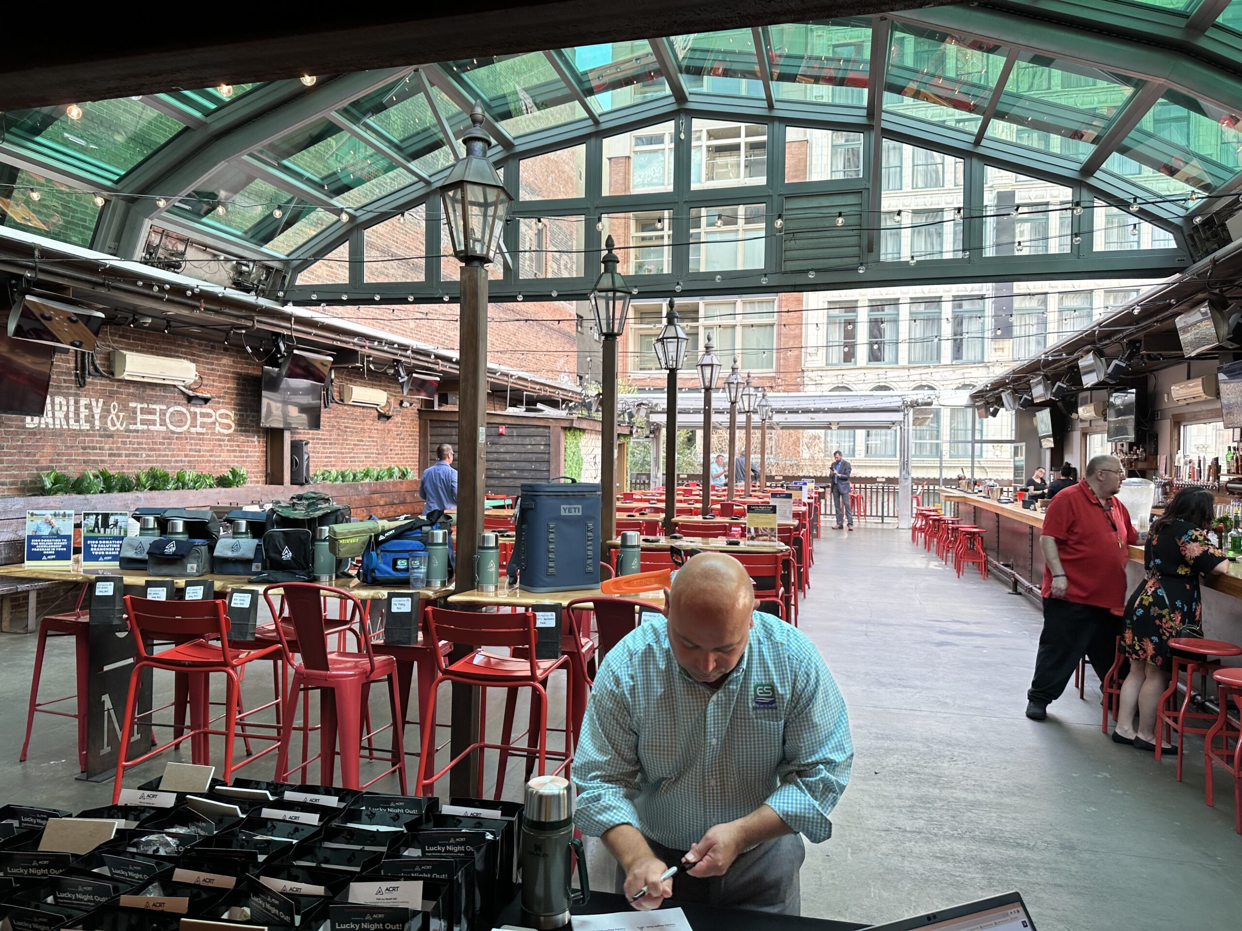 A man arranges items on a table at an indoor bar and event space with red chairs, a glass ceiling, and people mingling in the background.