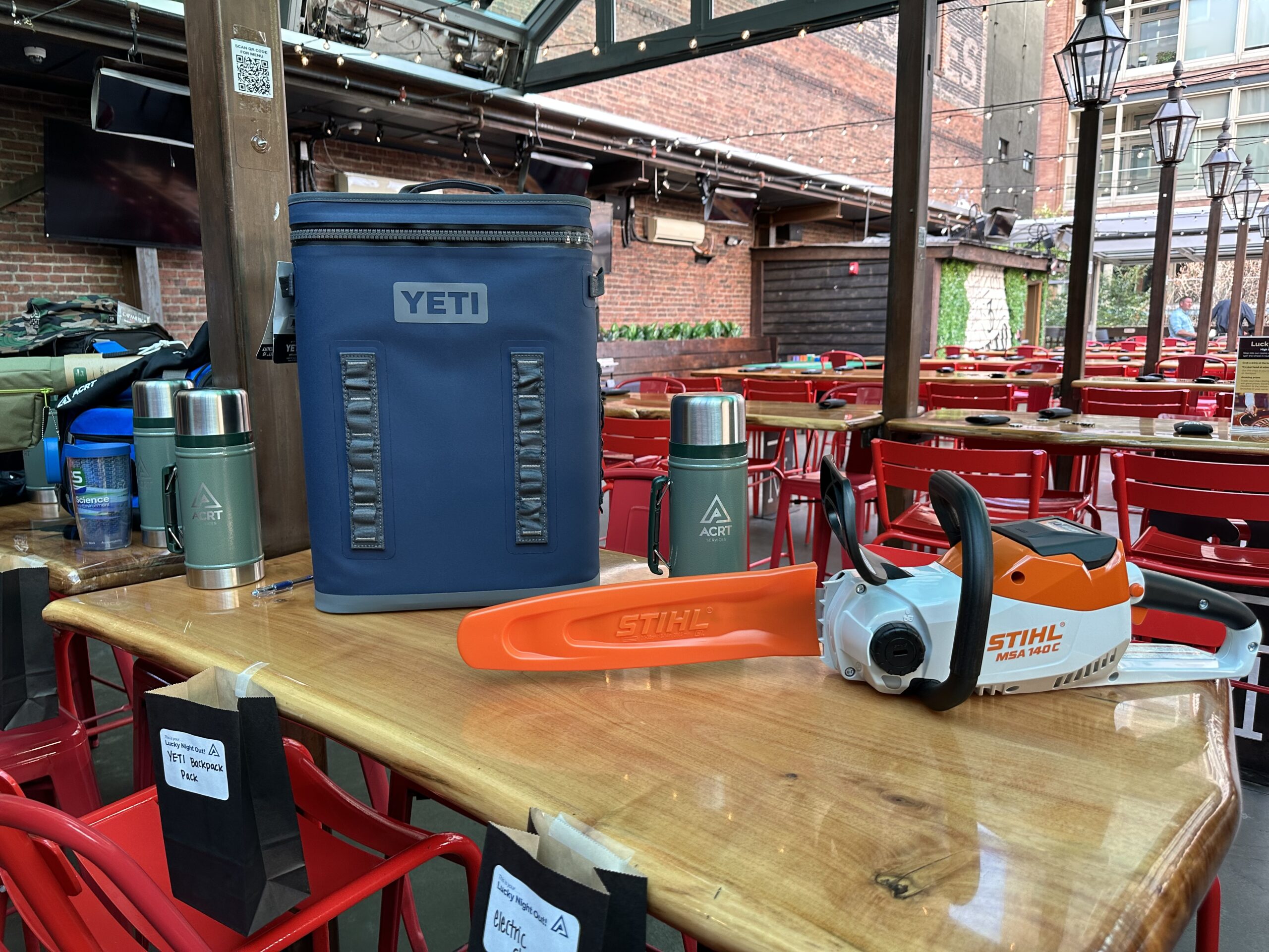 A YETI cooler, ACRT-branded thermoses, and a STIHL chainsaw are displayed on a table in a bar-style event space.