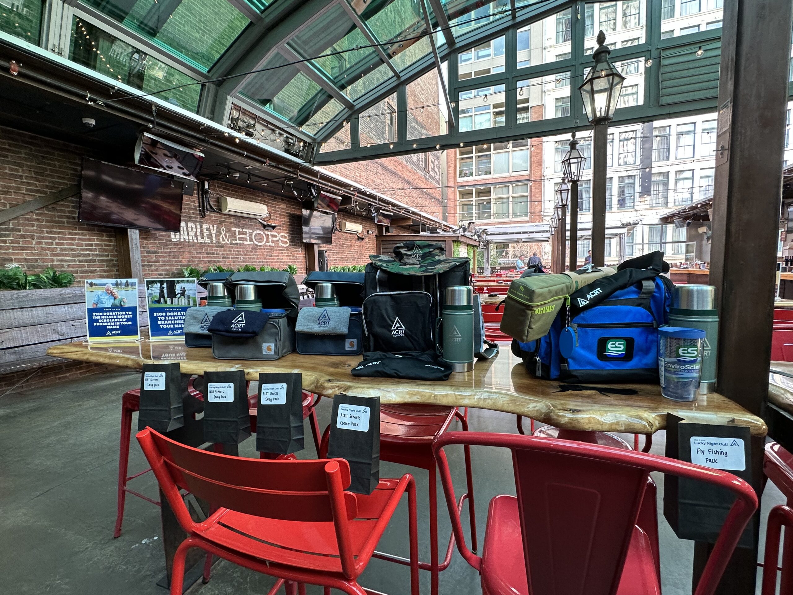 ACRT-branded bags, drinkware, and giveaway items are displayed on a table in a covered bar and event space with red chairs.