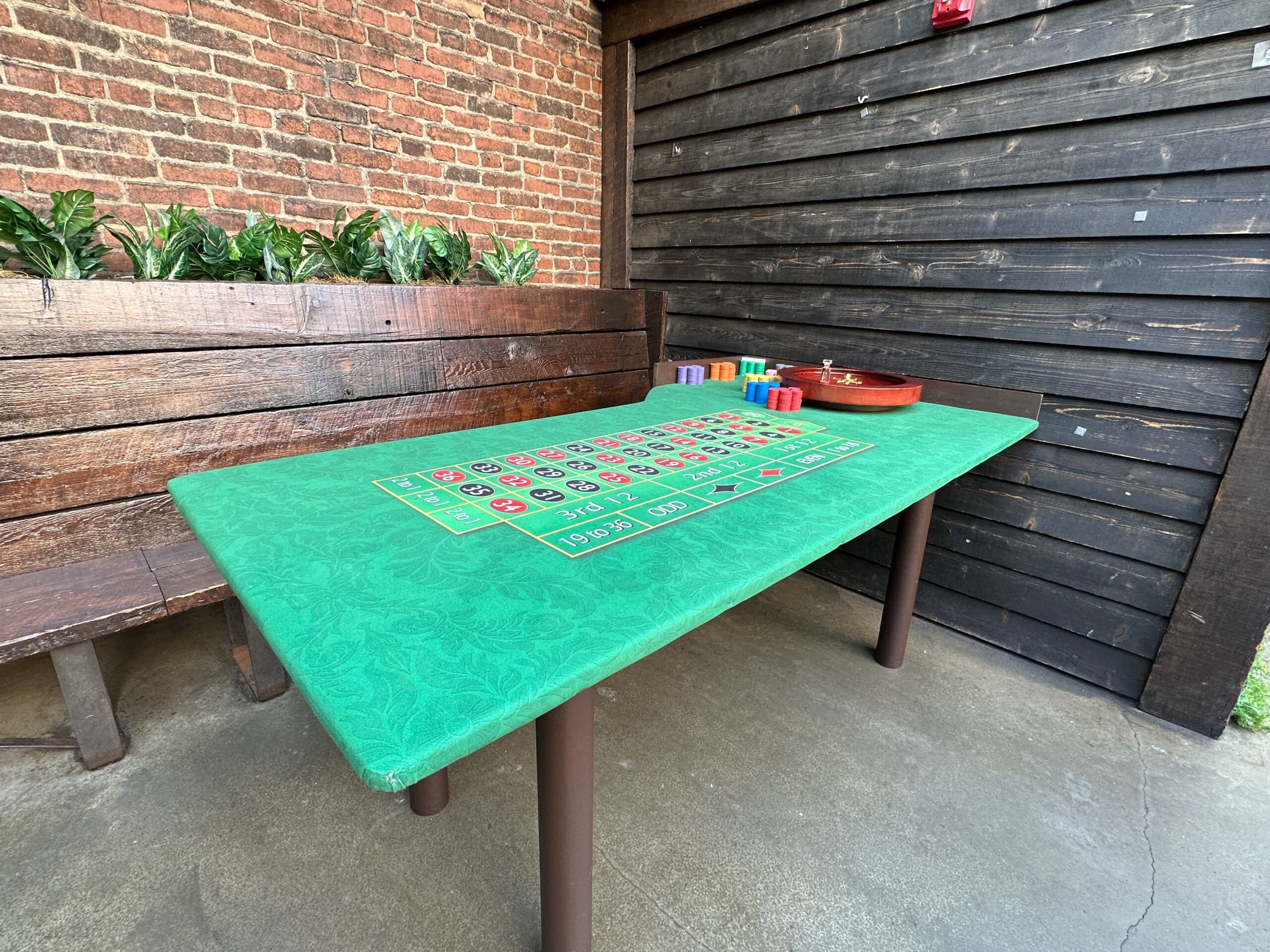 A roulette-style gaming table with chips and a tray is set up in a brick-walled patio area.