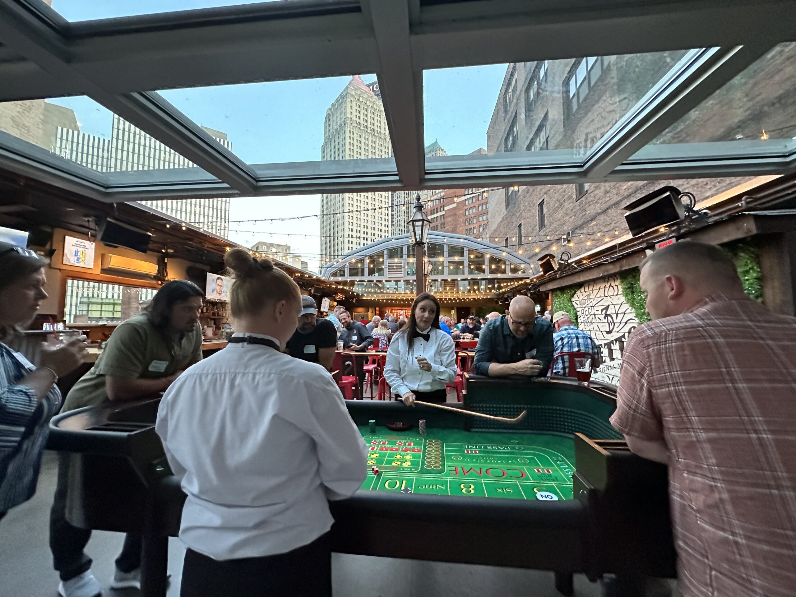 People standing around a craps table at an outdoor rooftop venue with city buildings in the background.
