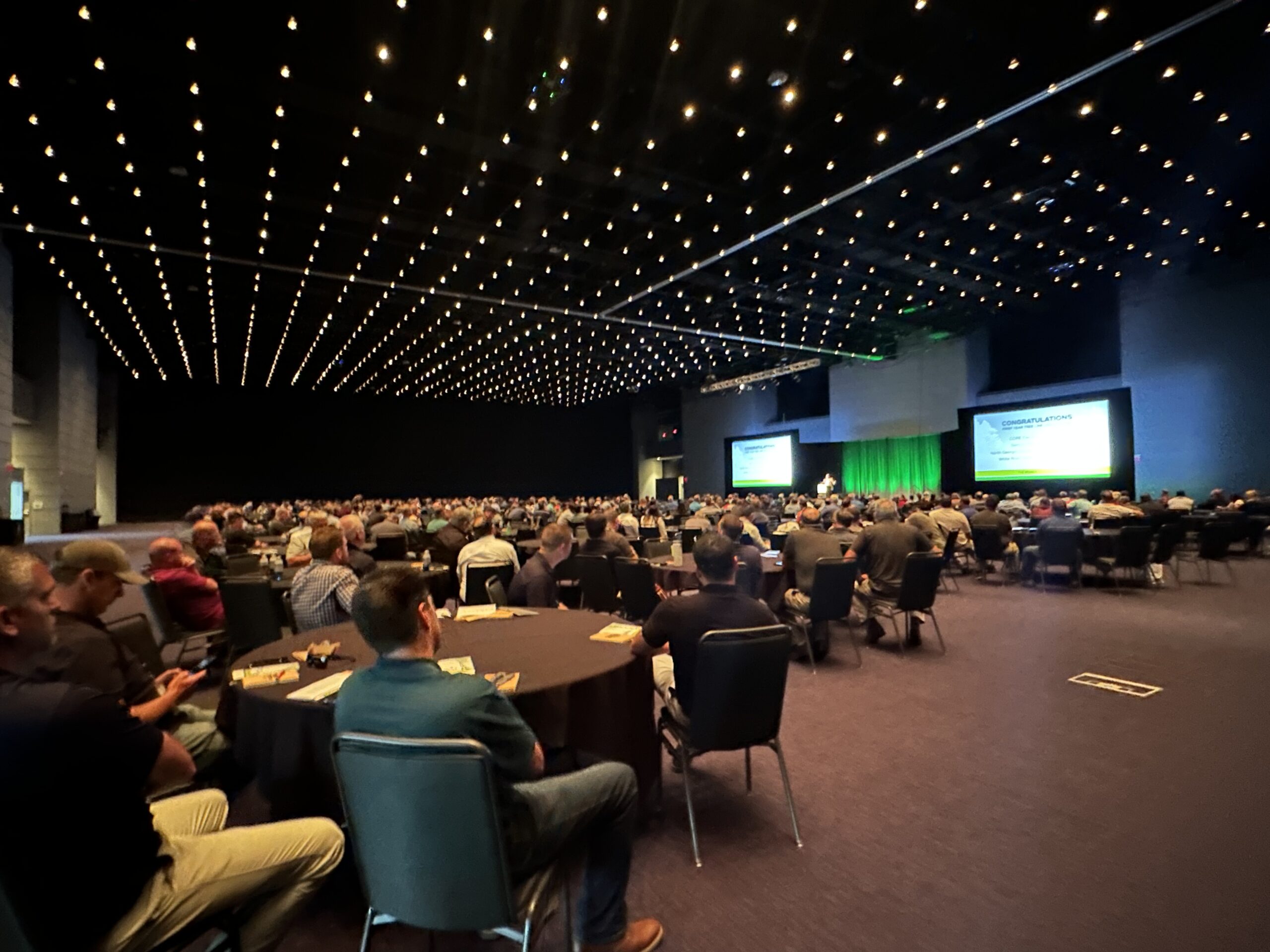 A large audience sits at round tables in a conference hall watching a presentation on a stage with green lighting.