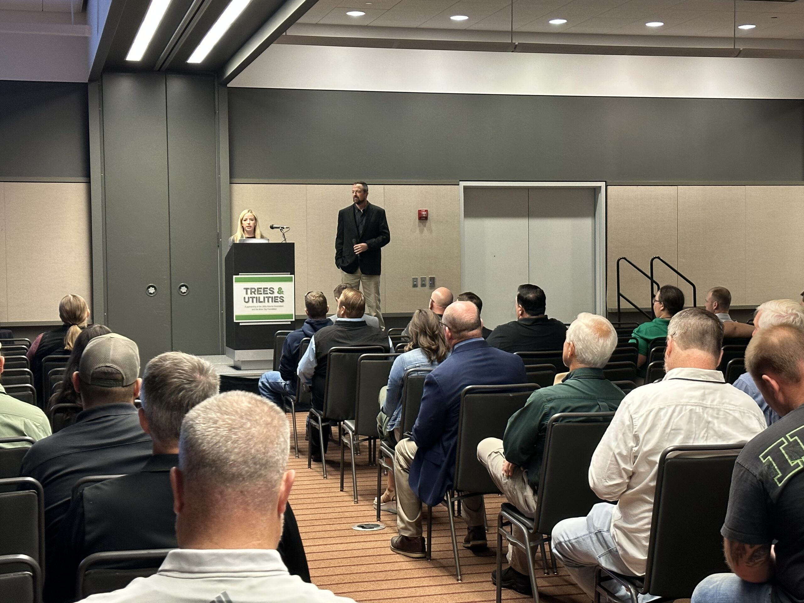 Two presenters speak at a podium labeled “Trees & Utilities” during a conference session as attendees listen.