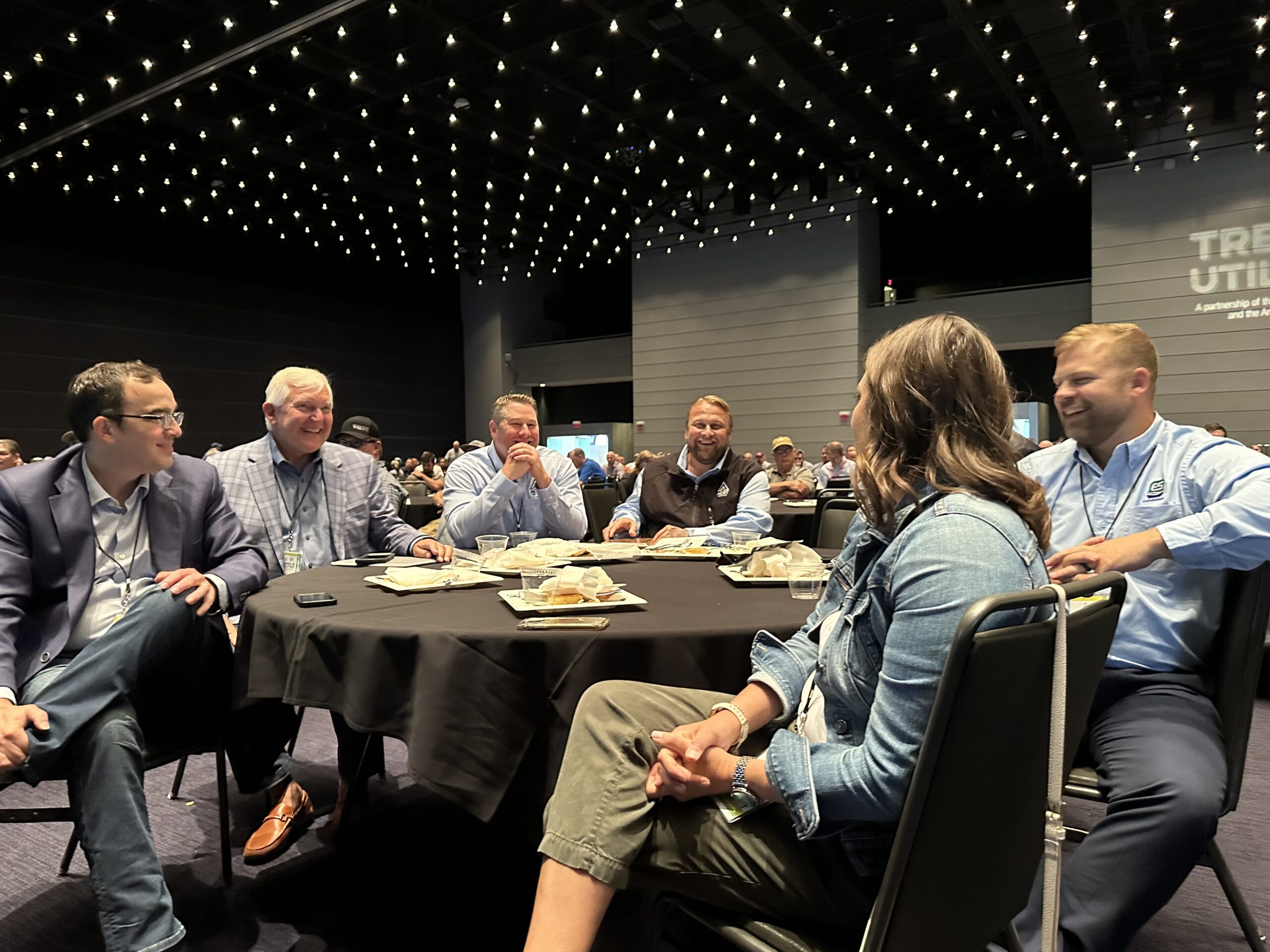Group of people seated around a table at an indoor conference event, talking and laughing.