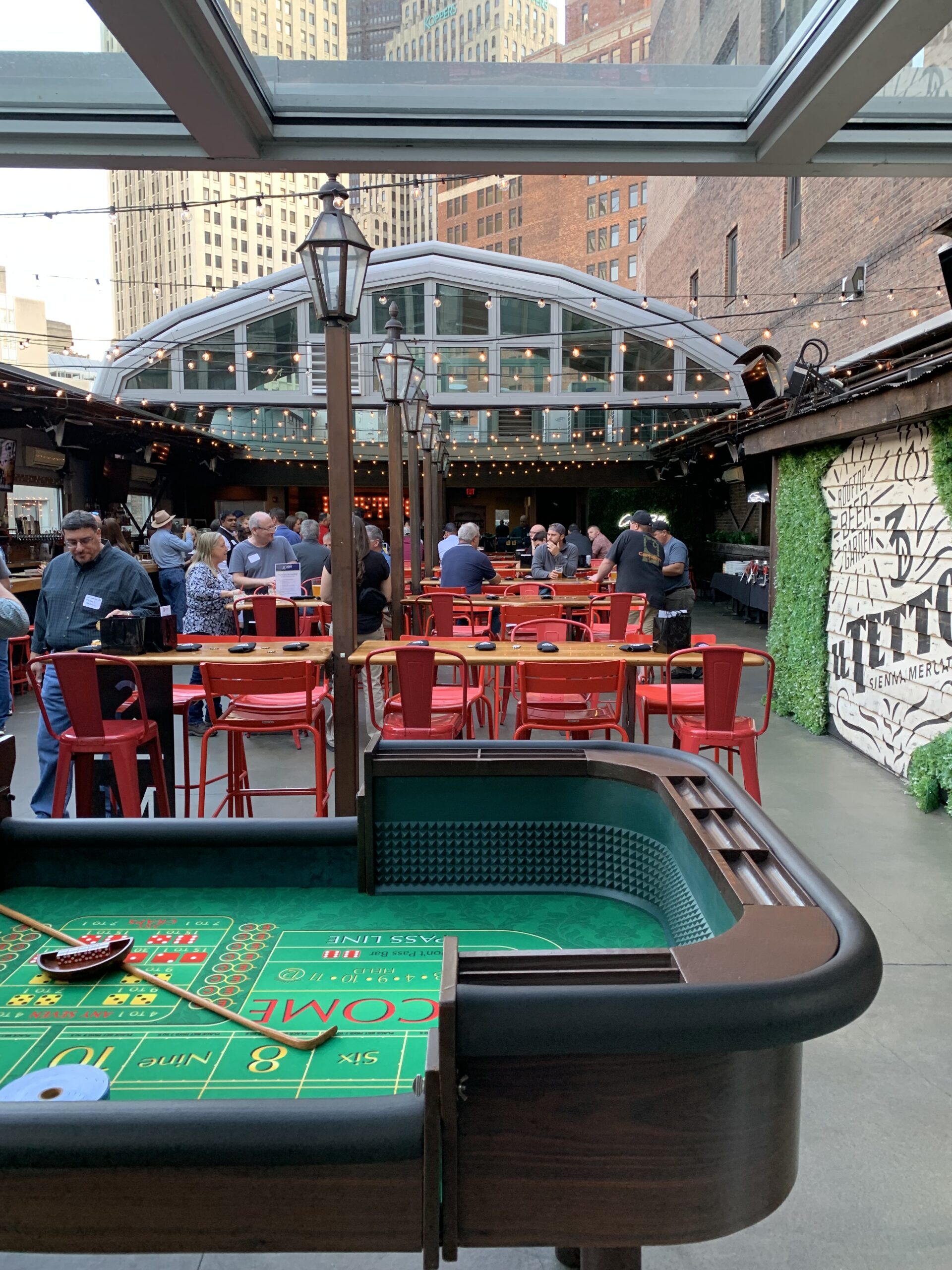 Outdoor restaurant or event patio with a craps table in the foreground and people seated and standing at tables in the background.