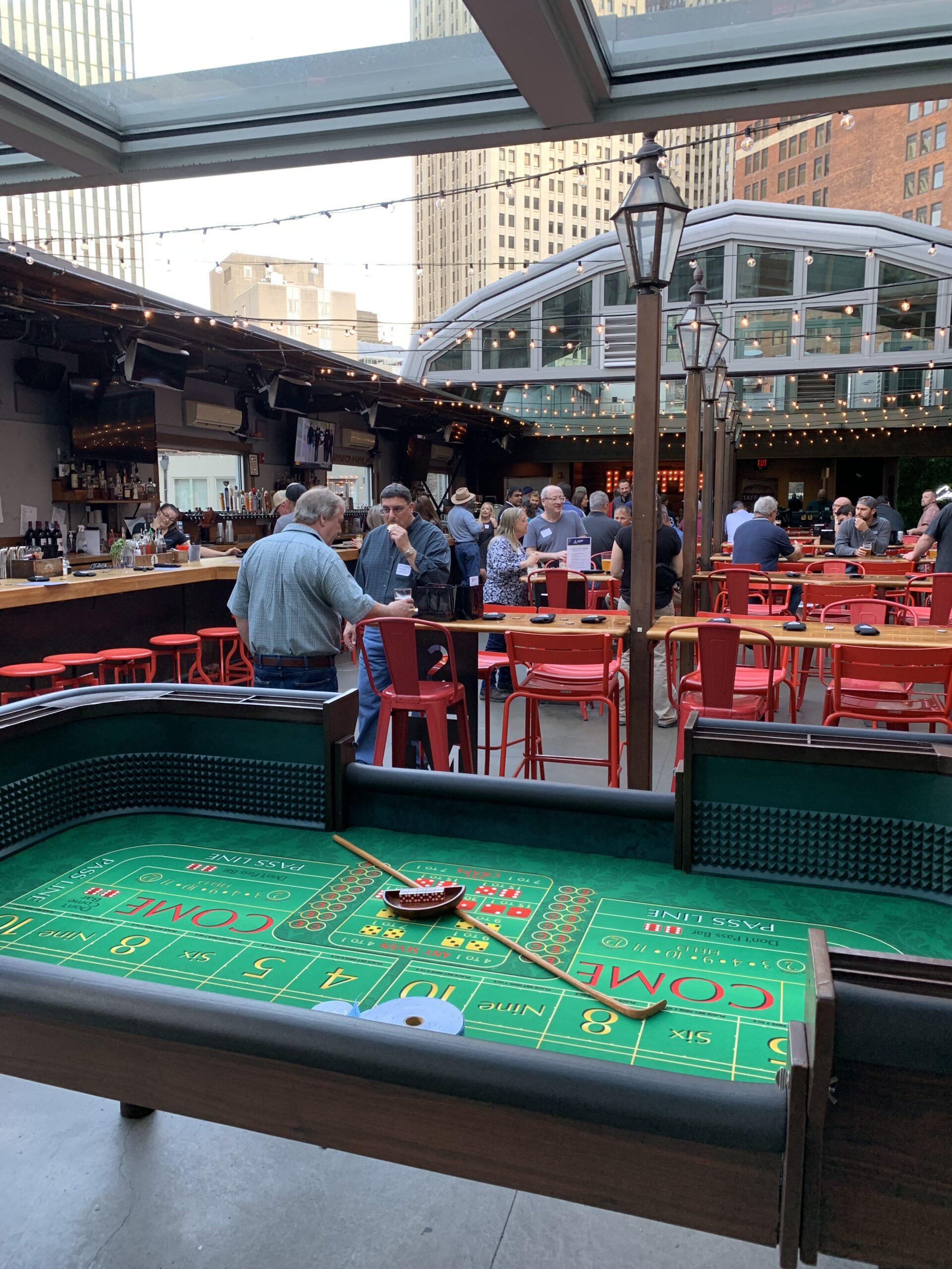 A craps-style gaming table sits in the foreground of a covered bar venue where attendees mingle.