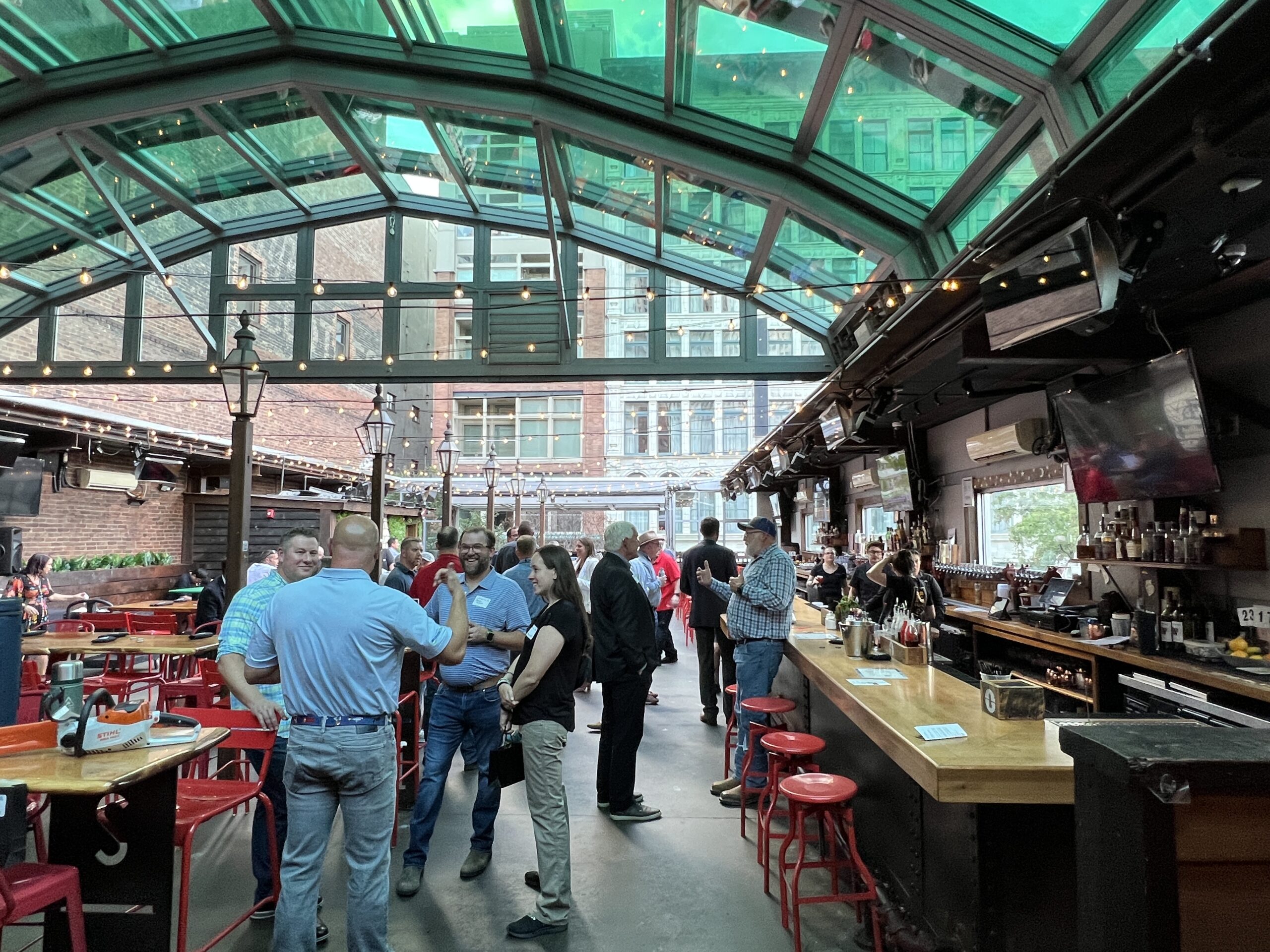People socializing in a covered bar event space with a glass roof and city buildings visible in the background.