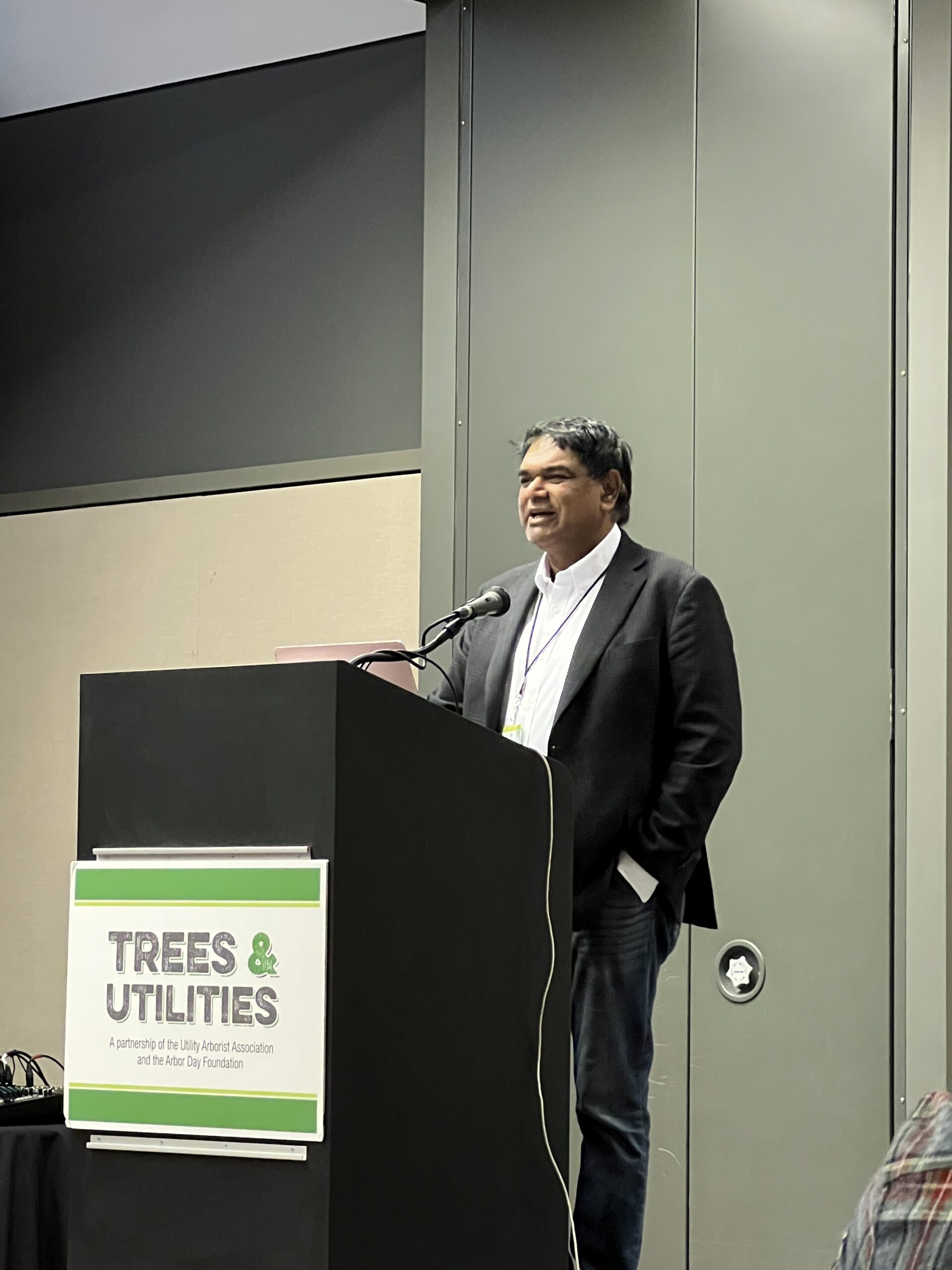 A man speaks at a podium labeled “Trees & Utilities” during a conference presentation.