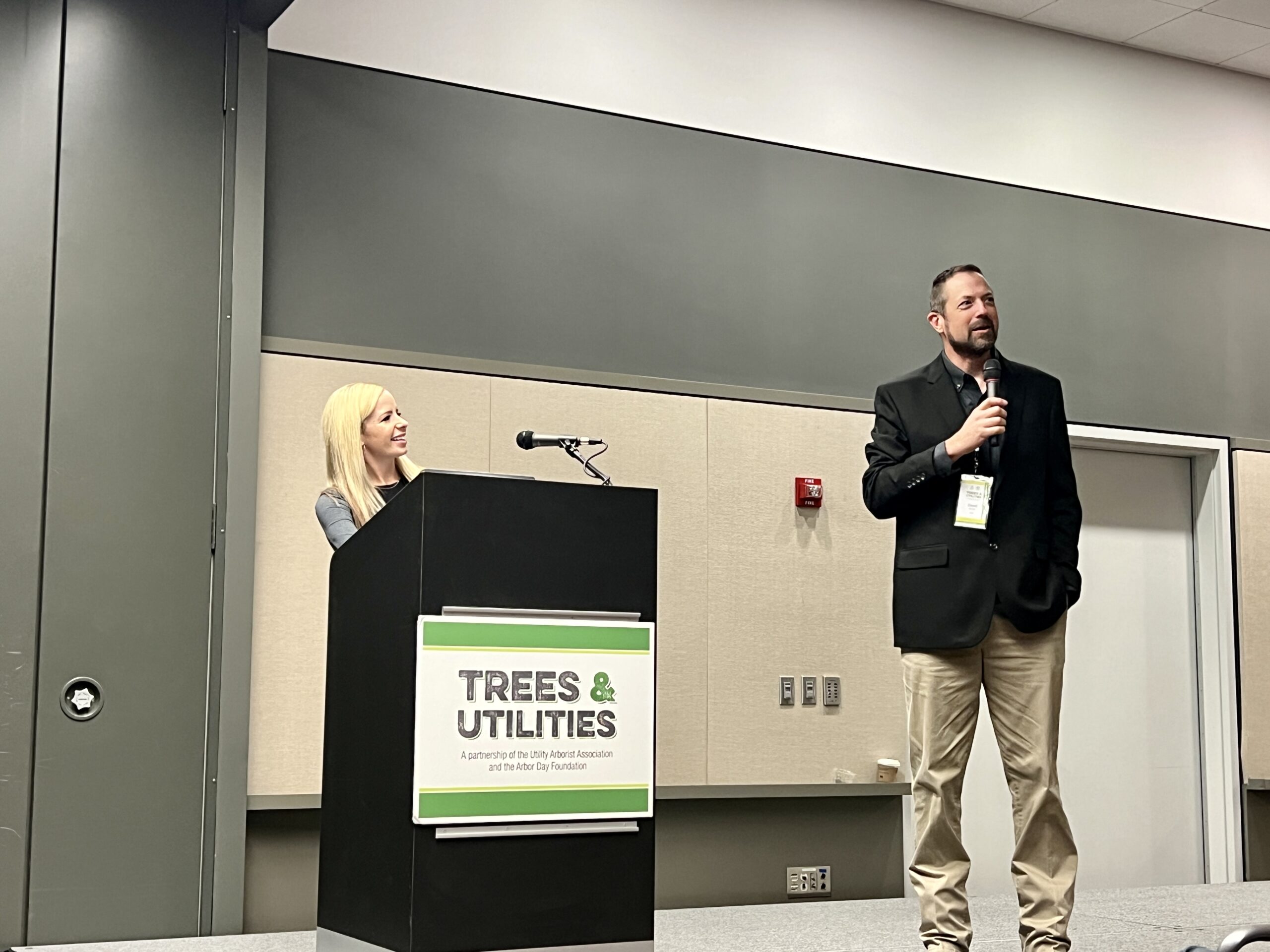 Two presenters speak on stage at a Trees & Utilities conference, one at a podium and one holding a microphone.