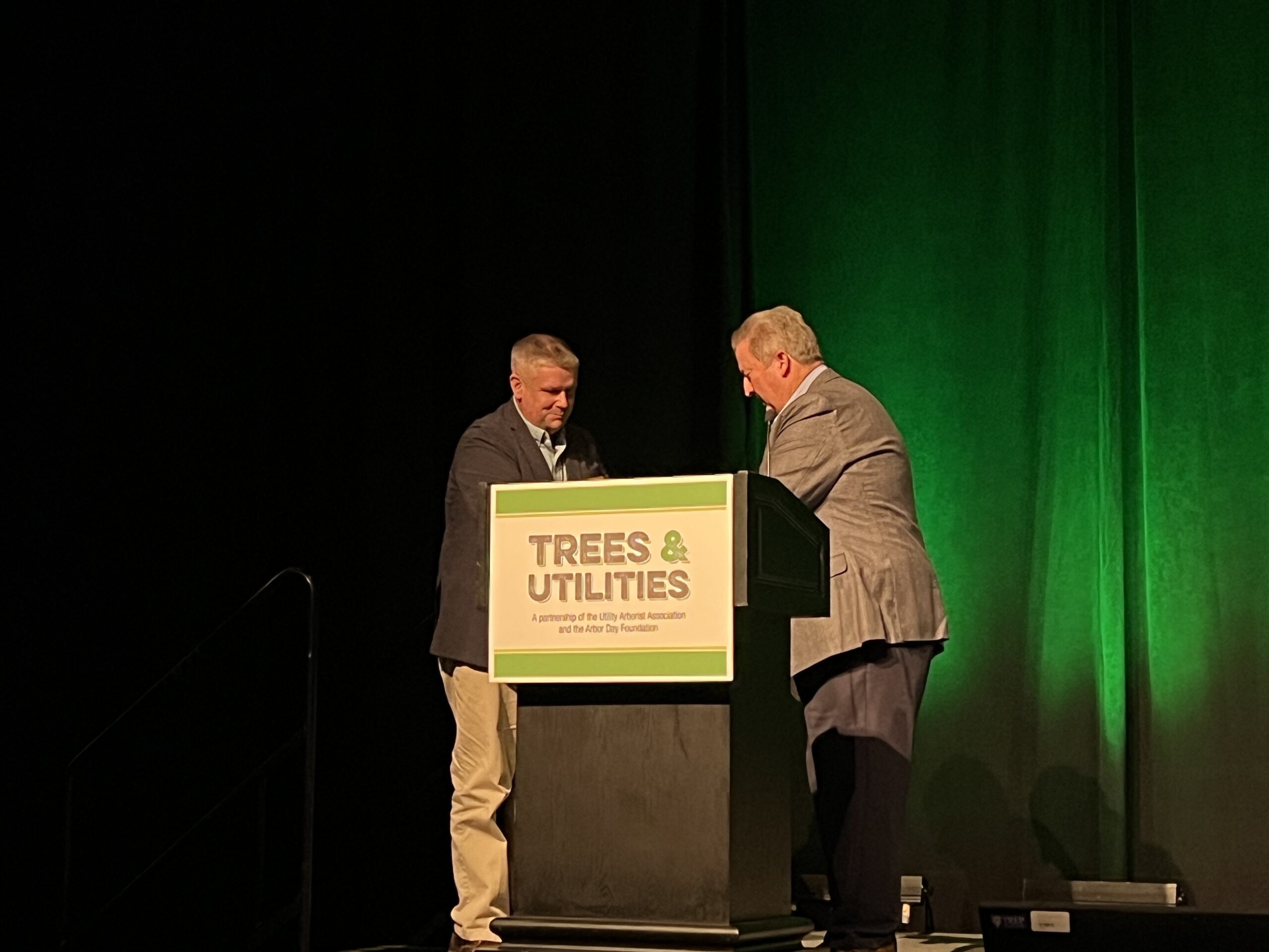 Two men stand at a podium labeled “Trees & Utilities” during a conference presentation, appearing to exchange or adjust materials.