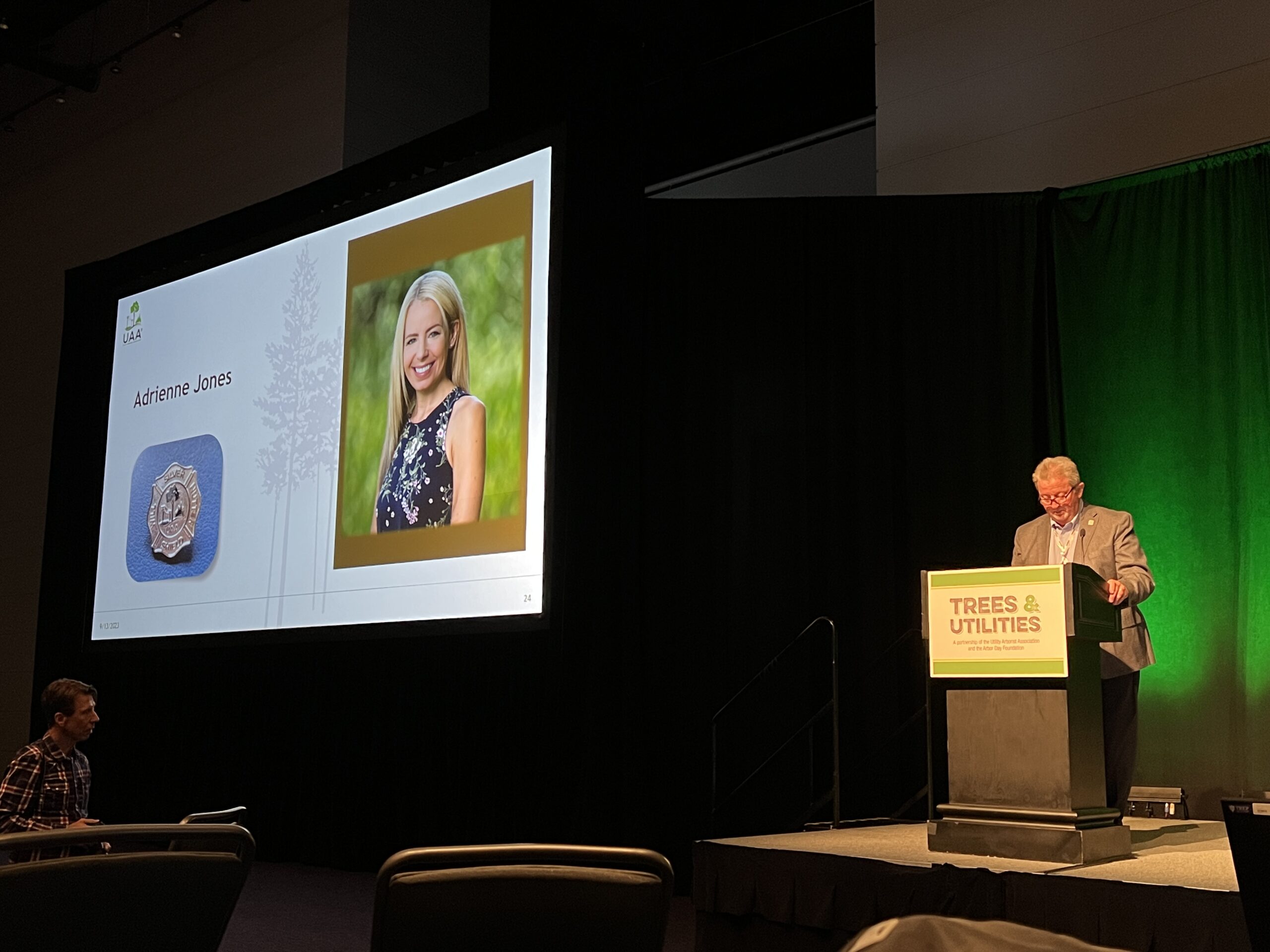 A speaker stands at a podium during a conference presentation while a slide showing a headshot and the name “Adrienne Jones” is displayed on a large screen.