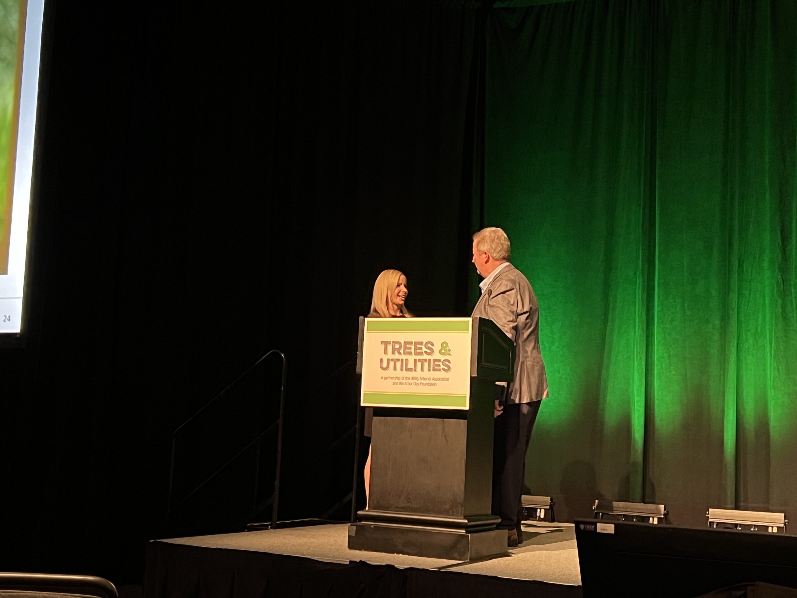 Two presenters stand at a podium labeled “Trees & Utilities” on a conference stage with green lighting.