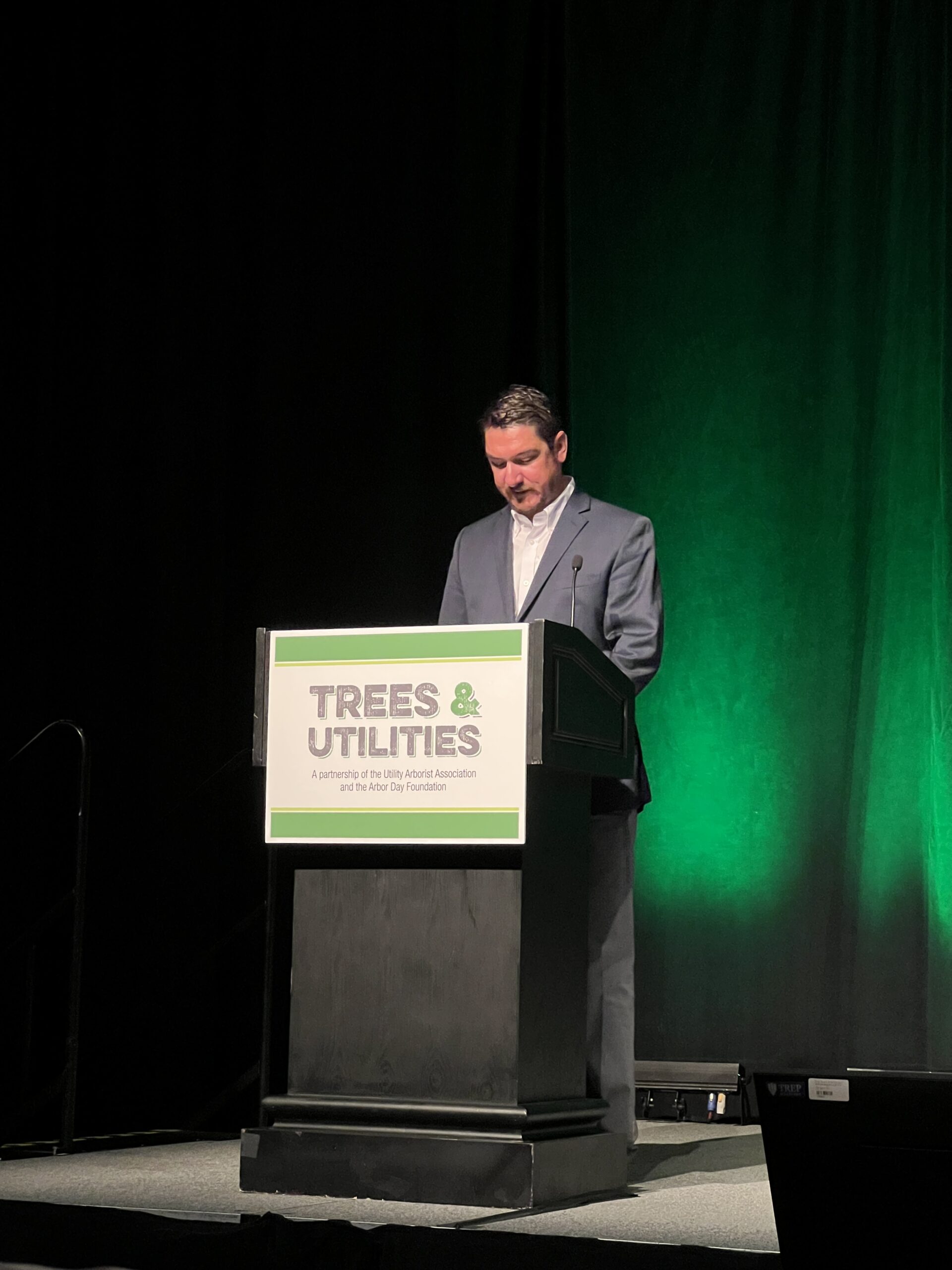 A speaker stands at a podium labeled “Trees & Utilities,” reading during a conference presentation with green stage lighting behind him.