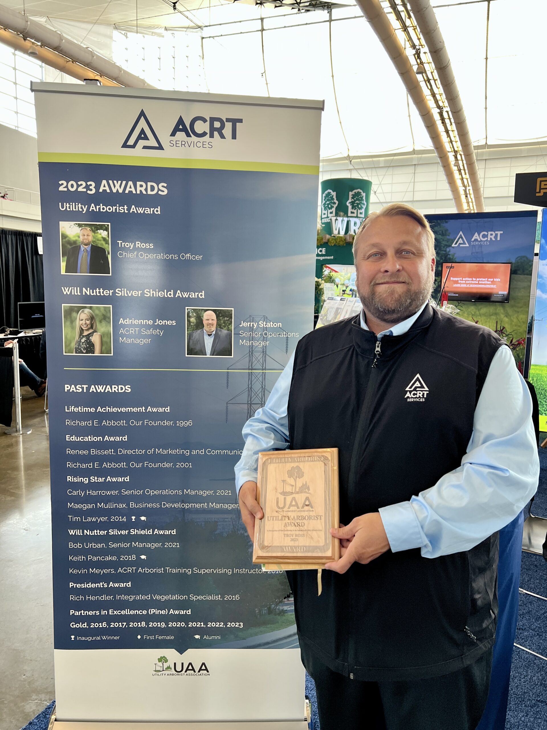 A man holds a UAA Utility Arborist Award plaque in front of an ACRT Services 2023 Awards display.