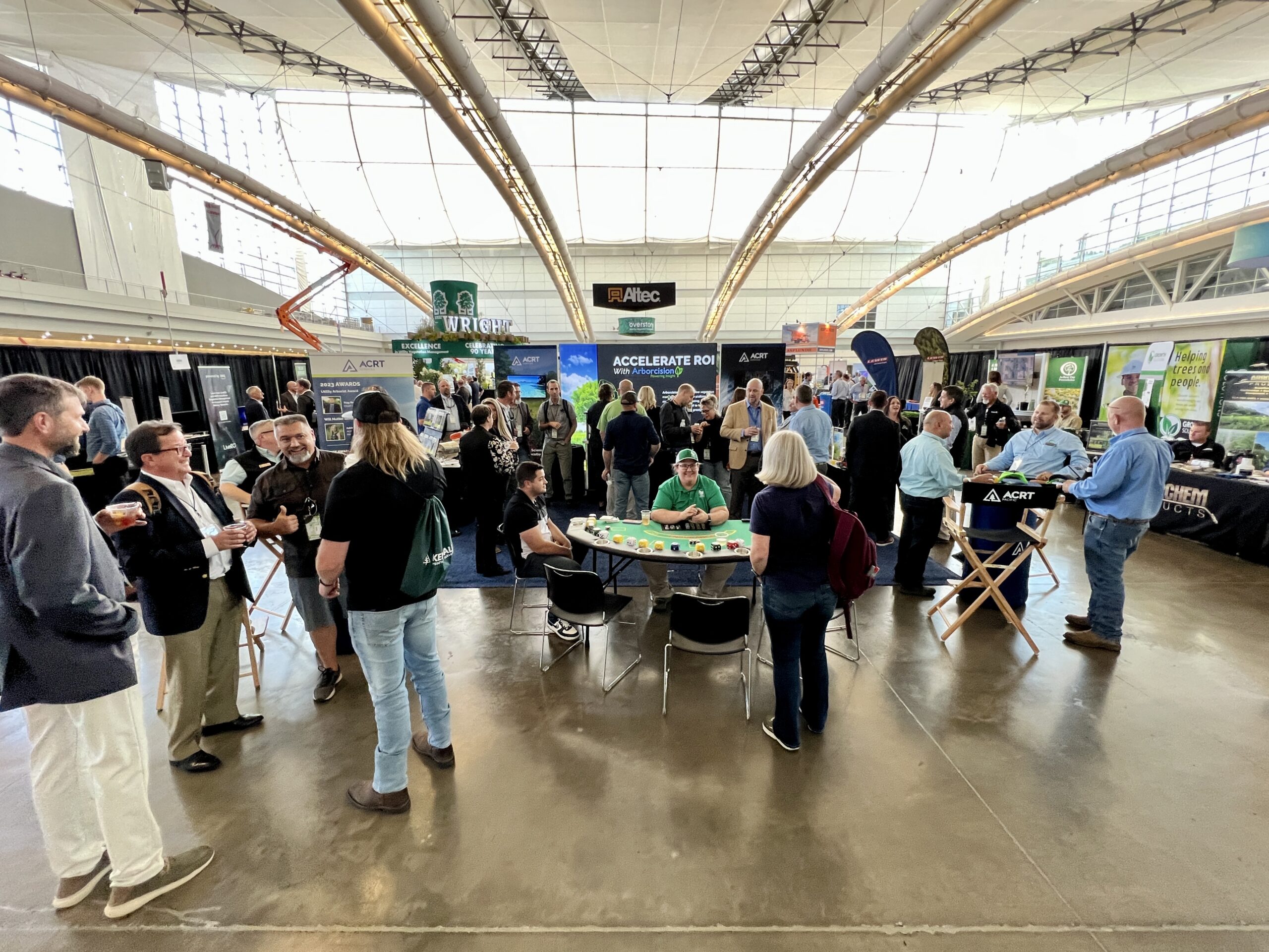Attendees gather and network at exhibitor booths inside a large, bright trade show hall.